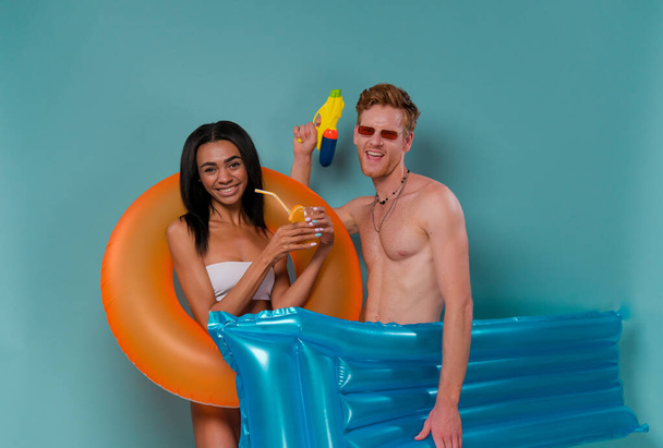 Pool party fun. Multiethnic Couple with inflatable rings having fun shooting with water guns, blue studio background. Summer vacation sea sun tan concept - Photo, Image