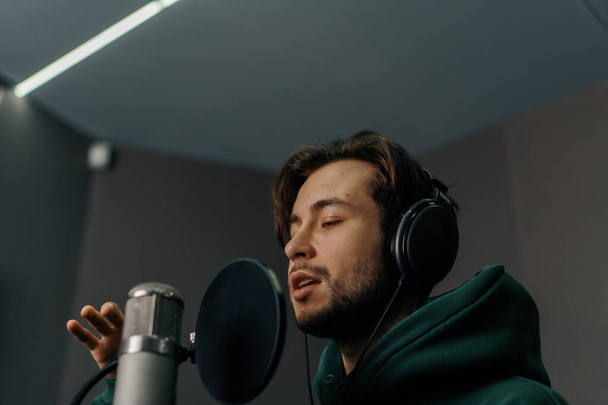 τραγουδιστής με ακουστικά και μικρόφωνο ηχογραφεί συναισθηματικά ένα νέο τραγούδι σε επαγγελματικό στούντιο ηχογράφησης - Φωτογραφία, εικόνα