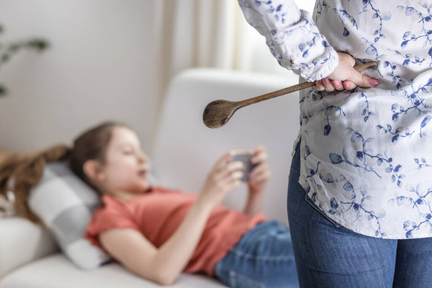 Adolescent girl ignores her mother standing upset with a wooden spoon next to her and keeps being on her mobile phone. - 写真・画像