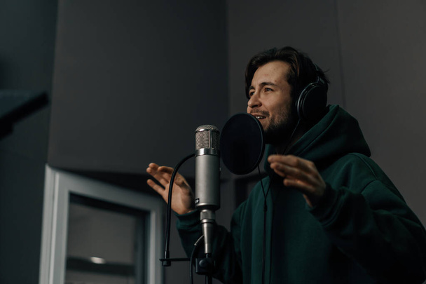 cantante con auriculares y micrófono grabando emocionalmente una nueva canción en un estudio de grabación profesional - Foto, Imagen