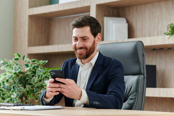 Businessman is smiling and typing a message on his smart phone in office. He is in a suit, workplace in modern and nice office. - Foto, afbeelding