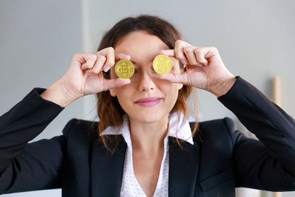 Cheerful young woman covering eyes with bitcoin coin currency in the office - Foto, afbeelding