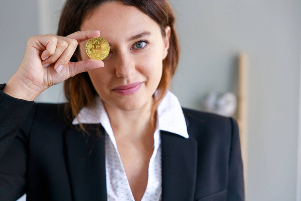 Smiling young woman covering the eye with bitcoin coin currency in the office. The focus is on the coin - Zdjęcie, obraz