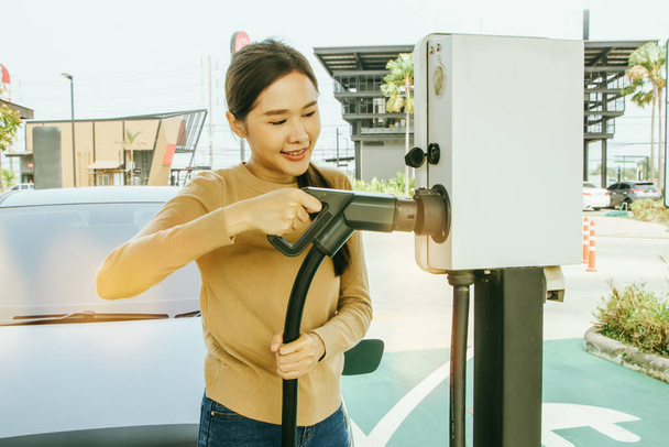 Retrato sonriente mujer asiática conduciendo un coche eléctrico que viaja a una estación de carga DC sosteniendo el cable de carga en un buen estado de ánimo: Conveniente estación de combustible eléctrico EV hace que las mujeres se sientan muy bien concepto. - Foto, imagen