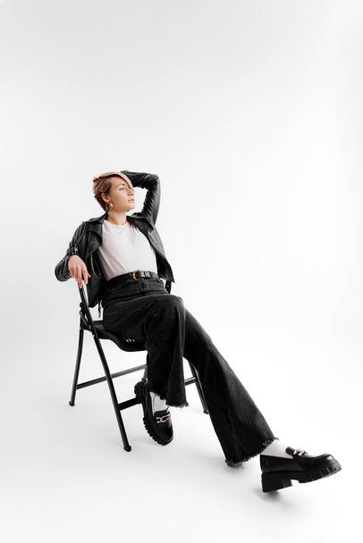 Full body of smiling young female in trendy black pants and leather jacket touching blond hair and relaxing on chair against white background - Valokuva, kuva
