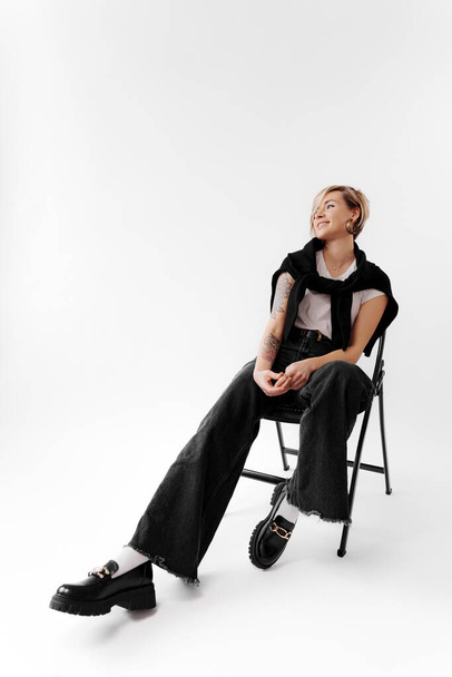 Full body of young female with leather jacket on shoulders and blond hair sitting on chair and relaxing against white background - Photo, Image