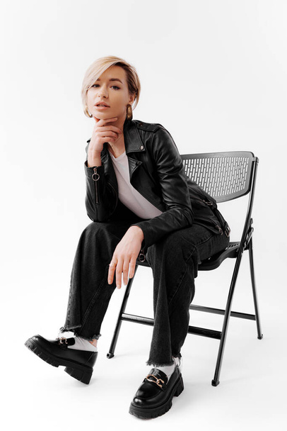 Full body of smiling young female in trendy black pants and leather jacket touching blond hair and relaxing on chair while looking at camera against white background - Photo, Image