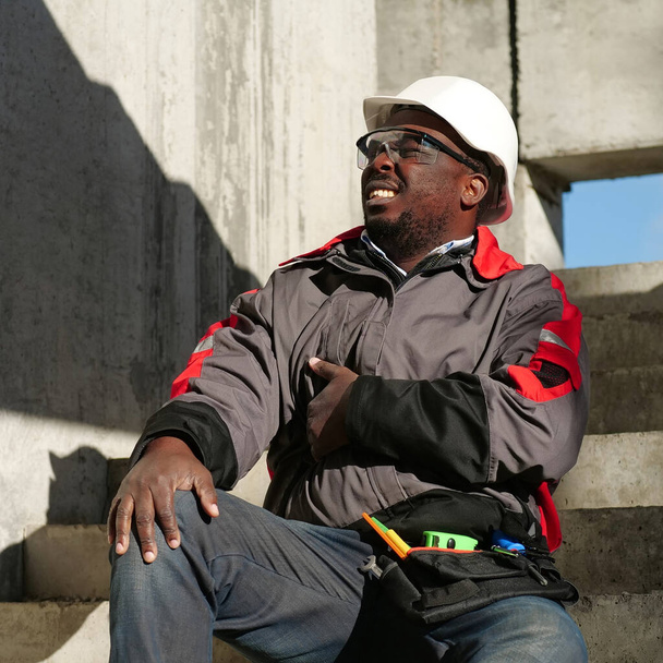 Afrikkalainen amerikkalainen työmies istuu portaissa rakennustyömaalla. Hän ei voi hyvin ja hänellä on vatsakipua, hän koskettaa vatsaansa - Valokuva, kuva