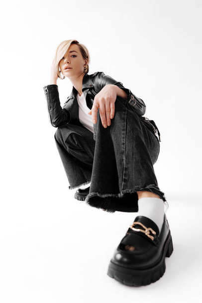 Full body of young female in leather jacket and boots sitting on haunches on floor and looking at camera while with hand touching hair against white background - 写真・画像
