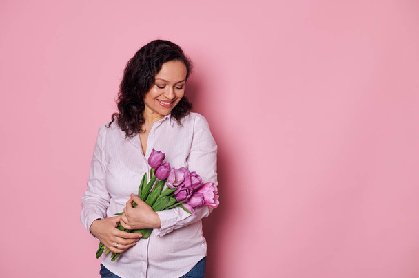 Beautiful young pregnant woman feeling happy, receiving a cute bouquet of purple tulips, smiles and expresses positive emotions, isolated over pink color background. Mothers Day and maternity concept - Photo, Image