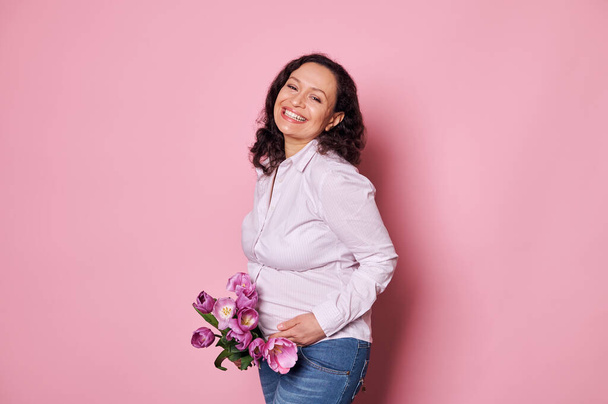 Šťastná kudrnatá multietnická těhotná žena v neformálním oblečení, usmívající se krásným zubatým úsměvem, pózující s kyticí tulipánů, izolovaná na růžovém pozadí. Mateřství. Šťastný Den matek - Fotografie, Obrázek