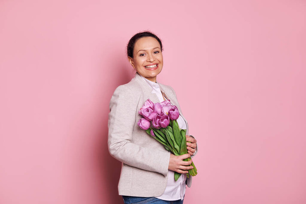 Zwangere multi-etnische vrouw glimlacht een tand glimlach kijken naar de camera, het vasthouden van een boeket van tulpen voor Womens Day of Moederdag, geïsoleerd over roze kleur achtergrond. Lente en moederschap concept - Foto, afbeelding