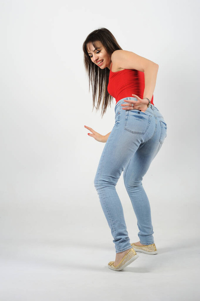 Young beautiful woman in a red shirt and blue jeans on a white background, with her back to the camera pulling something out of her pants pocket with one left hand( - Fotó, kép