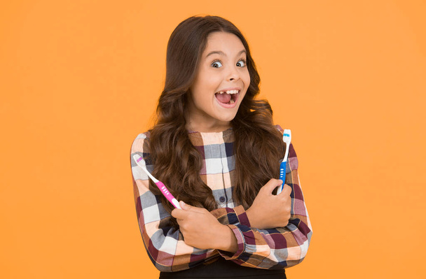 Teeth hygiene. Teeth whitening. Girl cute long hair holds toothbrushes yellow background. Child girl holds two toothbrushes. Child school girl smart kid happy face cares hygiene. Brush teeth concept. - Фото, изображение