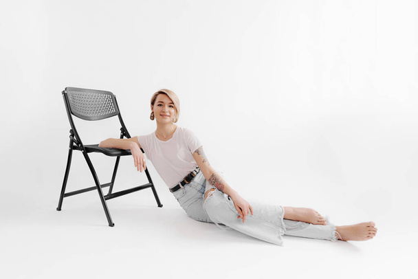 Вид збоку вдумливої жінки, що лежить з схрещеними ногами на підлозі і спирається на стілець і дивиться уві сні на білому тлі
 - Фото, зображення
