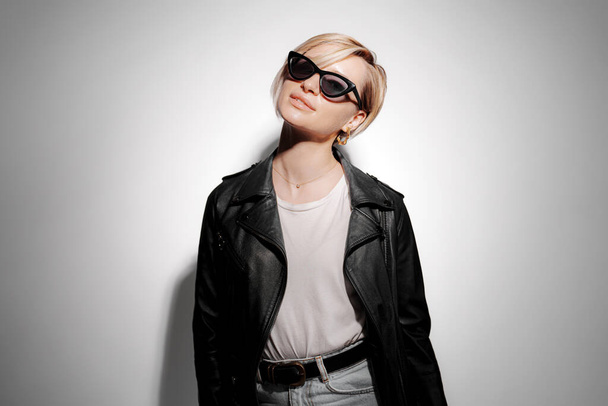 Confident young female in trendy black leather jacket and sunglasses standing against gray background in light with shadow and looking at camera - Foto, Imagen