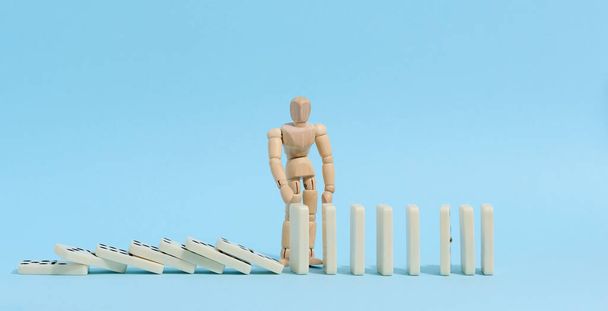 Ein Holzpuppenspielzeug hält einen fallenden Dominostein auf blauem Hintergrund zurück, der das Konzept einer starken Persönlichkeit repräsentiert. - Foto, Bild