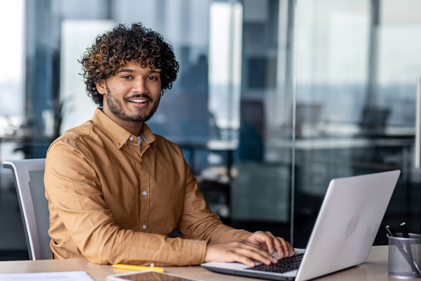 Portrait of young successful Indian man at workplace inside office, businessman smiling and looking at camera, man at work using laptop, programmer with curly hair coding software. - 写真・画像