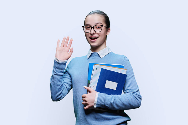 Portrait d'une lycéenne souriante avec des cahiers de notes dans les mains sur fond de studio blanc. Smart amical adolescent femme agitant à la caméra. Adolescence, éducation, concept d'étude - Photo, image