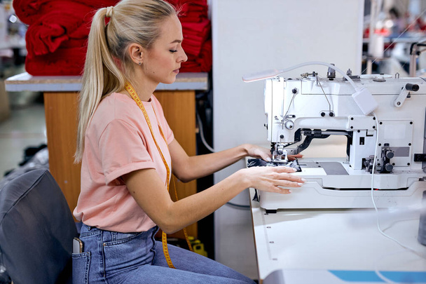 focused blonde caucasian female seamstress sits using modern sewing machine. Dressmaker works in textile workshop. Tailoring, sewing as a small business or hobby concept. sewing process - Zdjęcie, obraz