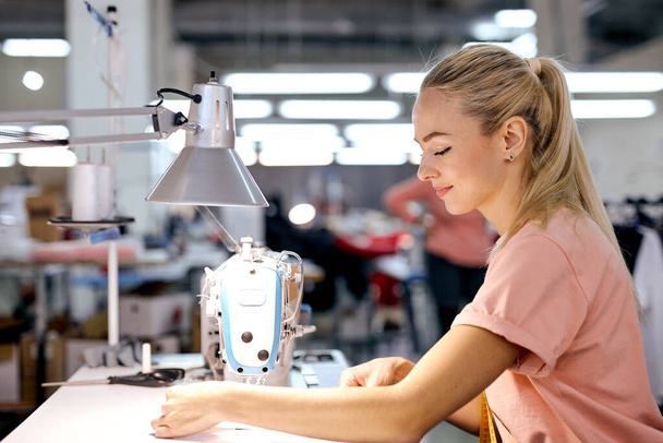 young nice caucaian blonde textile factory worker on production line using modern sewing machine, at workshop. hardworking woman engaged in sewing or tailoring, dressmaking concept - Фото, изображение