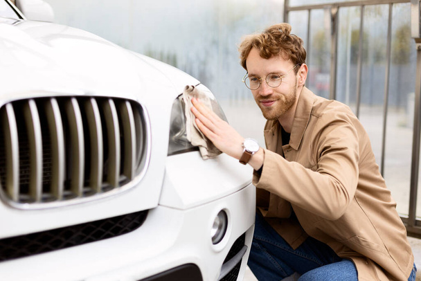 Detailní záběr usměvavého, atraktivního muže, který otírá reflektory auta mikrovláknitou látkou. Koncept samoobslužnosti, mytí aut - Fotografie, Obrázek