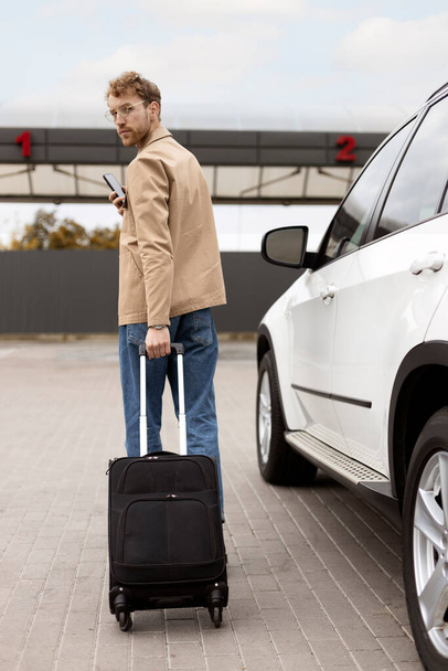 Achteraanzicht, serieuze man die met koffer loopt, smartphone vasthoudt, in de buurt van een auto. Concept van reizen, weg, autorit - Foto, afbeelding
