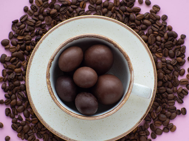 Flat lay composition with small chocolate easter eggs in cup surrounded by coffee roasted beans. Easter coffee concept. Mug full of sweet brown eggs on a plate with coffee beans on a pink background. - Foto, Bild
