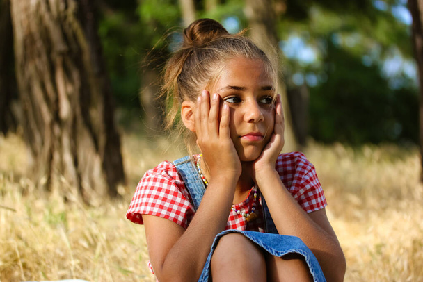 Een mooi meisje van 10-12 jaar oud zit in de zomer op een open plek in het bos. Een kind verveelt zich tijdens de zomervakantie. Problemen in een gezin, kinderpsychologie, saai concept.  - Foto, afbeelding