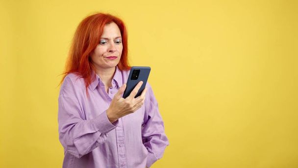 Worried redheaded mature woman using a mobile phone in studio with yellow background - Φωτογραφία, εικόνα