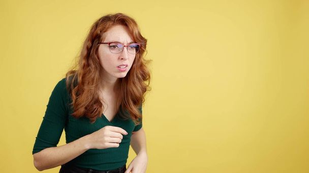 Rudowłosa kobieta w okularach szpieguje z ciekawością w studiu z żółtym tłem - Zdjęcie, obraz