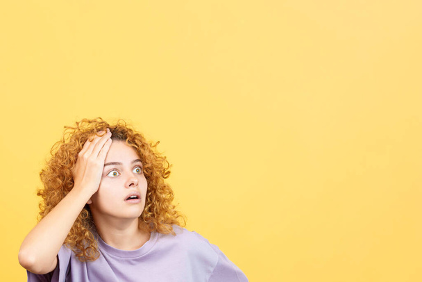 Studio image with yellow background of a woman with curly hair gesturing with the hand on head expressing despair - Foto, afbeelding