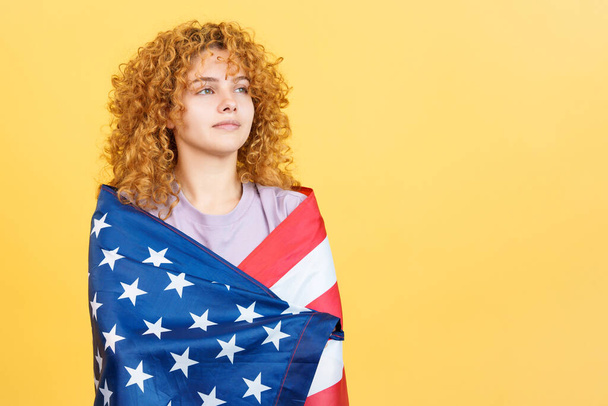 Studiobild mit gelbem Hintergrund einer Schönheit und abgelenkten ernsten Frau mit lockigem Haar, die sich mit einer nordamerikanischen Nationalflagge umwickelt - Foto, Bild