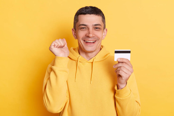 Online shopping. Banking paying. Delighted cheerful man wearing casual hoodie earning big sum of money clenched fist celebrating succes posing isolated over yellow background. - Photo, Image