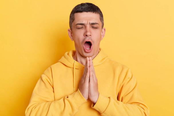 Funny man wearing casual hoodie meditating to calm down saying ohm standing with open mouth keeps palms together posing isolated over yellow background. - Foto, Bild