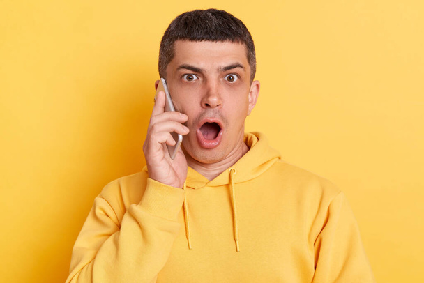Scared man wearing casual hoodie having conversation on cell phone talking with friends keeps mouth open hearing news posing isolated over yellow background. - Fotó, kép