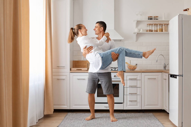Young couple posing in modern kitchen smiling husband holding his wife enjoying gentle moment having fun wearing white shirts and jeans. - Photo, Image