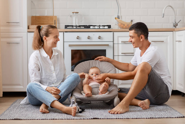 Young family with little child sitting on floor in new modern design kitchen, parents playing with their baby in rocking chair expressing happiness. - Foto, immagini
