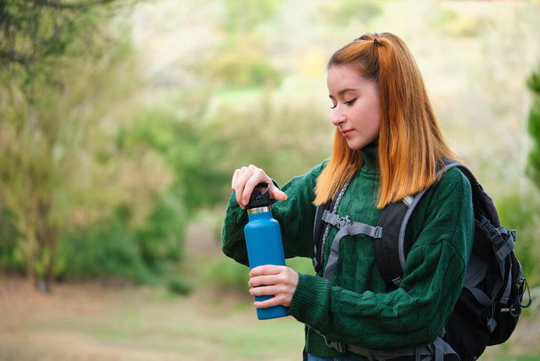 Χαμογελαστή πεζοπόρος κοκκινομάλλα νεαρή γυναίκα ανοίγει ένα μπουκάλι νερό στο βουνό φορώντας ένα σακίδιο. Έννοια πεζοπορίας. - Φωτογραφία, εικόνα