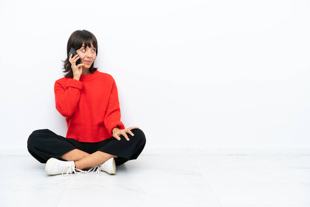 Jeune femme métisse assise sur le sol isolée sur fond blanc gardant une conversation avec le téléphone portable avec quelqu'un
 - Photo, image