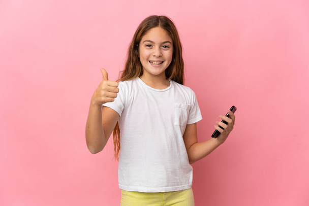 Dziecko nad odizolowanym różowym tle za pomocą telefonu komórkowego podczas wykonywania kciuki w górę - Zdjęcie, obraz