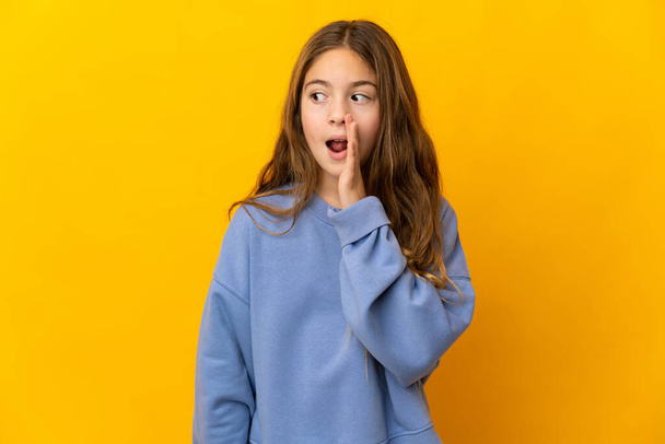 Child over isolated yellow background whispering something with surprise gesture while looking to the side - Photo, Image