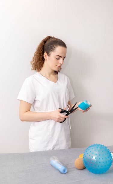 Rehabilitation Specialist, Physical Therapist With Rehab Tools Cuts Elastic Kinesiology Tape With Scissors In Therapeutic Room. Vertical plane. High quality photo - Photo, Image