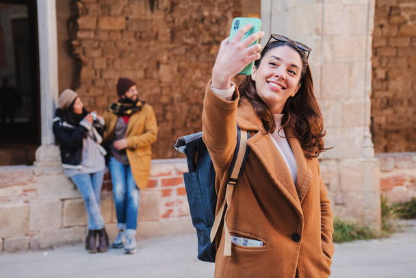 En primer plano una joven mujer caucásica tomando un retrato de selfie con un teléfono móvil al aire libre, en el fondo una pareja hablando. mujer turística aislada disparando una foto con abrigo y mochila. Alto. - Foto, Imagen