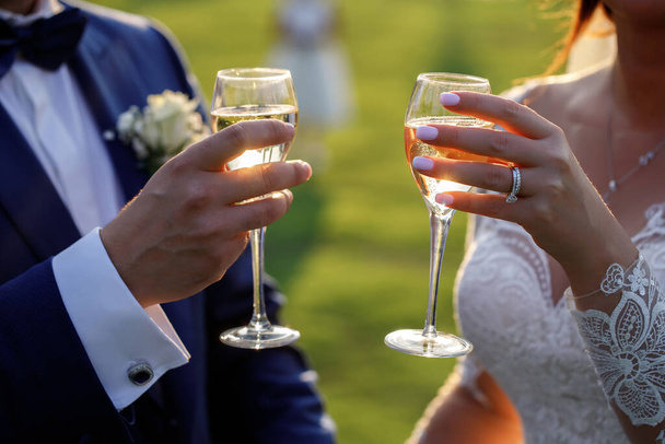 O noivo em um terno clássico azul e a noiva em um vestido de casamento branco, laço, com belos abotoaduras, um grande anel, bebe a champanha de copos no pôr do sol. Mãos sem rostos close-up. - Foto, Imagem