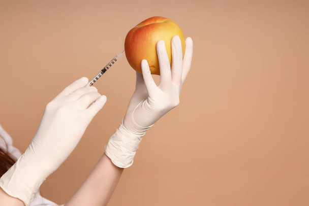 the hand of a young girl in medical latex gloves makes an injection with a filler syringe into a large red apple on a brown background close-up. cosmetic procedure for cell rejuvenation - Foto, Imagen