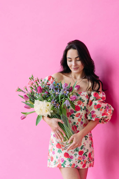 Young happy woman stands and holds vase with flowers bouquet in her hands against pink background. Mothers day, birthday, anniversary, celebration, holiday, greeting, festive event. - Photo, Image