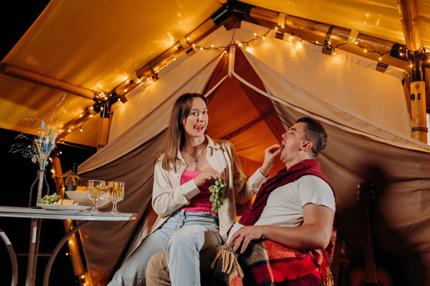 Happy lovely couple relaxing in glamping on summer evening and eating grapes near cozy bonfire. Luxury camping tent for outdoor recreation and recreation. Lifestyle concept - Zdjęcie, obraz