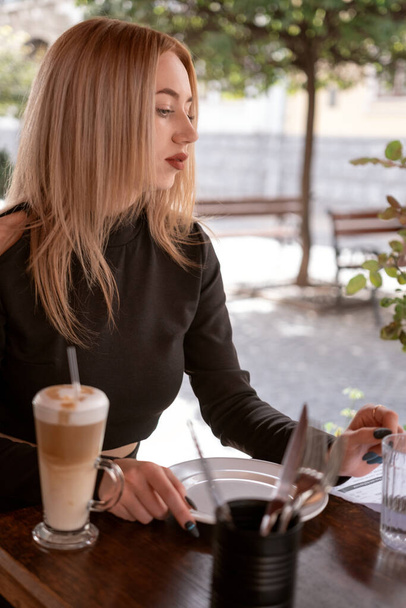 Retrato de chica rubia atractiva en la cafetería. Joven hermosa mujer espera su pedido en la cafetería y bebe café con leche - Foto, imagen