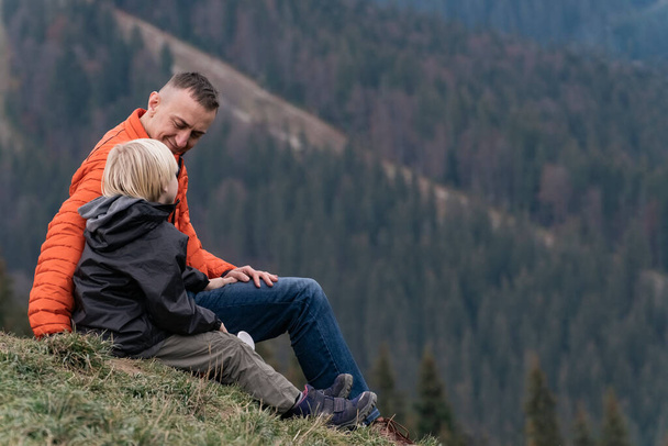 Отец и сын сидят на склоне холма на фоне леса. Выходные прогулки в горах. Пешие прогулки и походы с детьми - Фото, изображение